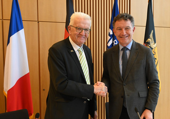 Grenzraumkonferenz Baden-Württemberg und Grand Est vertiefen Zusammenarbeit Ministerpräsident Winfried Kretschmann (links) und Franck Leroy (rechts), Präsident der Region Grand Est, stehen vor Fahnen und geben sich die Hand.
