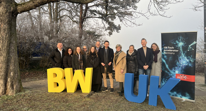 Eine Gruppe Menschen steht auf einer Wiese. Im Hintergrund ist ein Baum zu sehen. Vor der Gruppe stehen gelbe und blaue Buchstaben: BW und UK.
