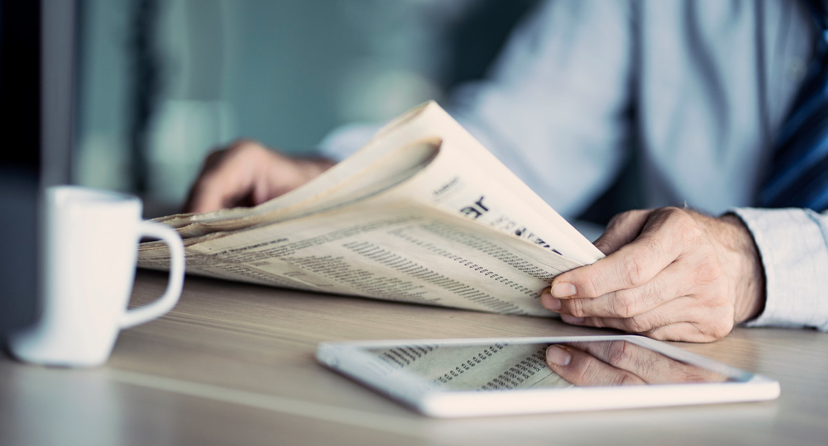 Eine Person, die nur im Anschnitt zu sehen ist, hält eine Zeitung in der Hand.
