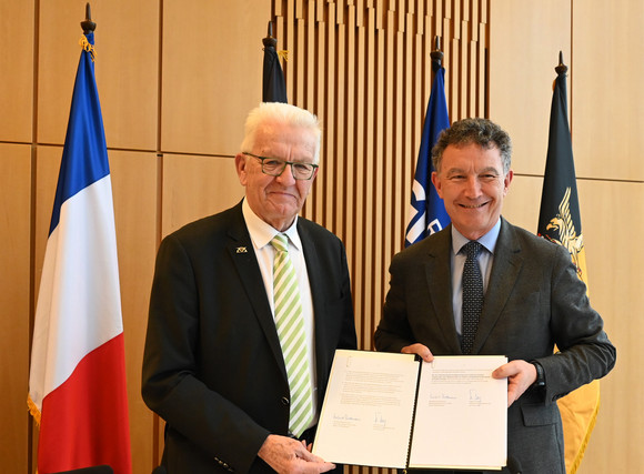 Grenzraumkonferenz Baden-Württemberg und Grand Est vertiefen Zusammenarbeit Ministerpräsident Winfried Kretschmann (links) und Franck Leroy (rechts), Präsident der Region Grand Est, stehen vor Fahnen und haben die unterzeichneten Positionspapiere in der Hand.