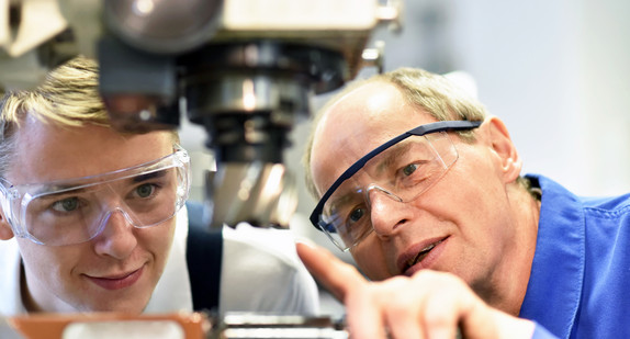Ein Mann mit einer Schutzbrille zeigt auf eine Maschine. Neben ihm ein junger Mann ebenfalls mit einer Schutzbrille der darauf schaut.