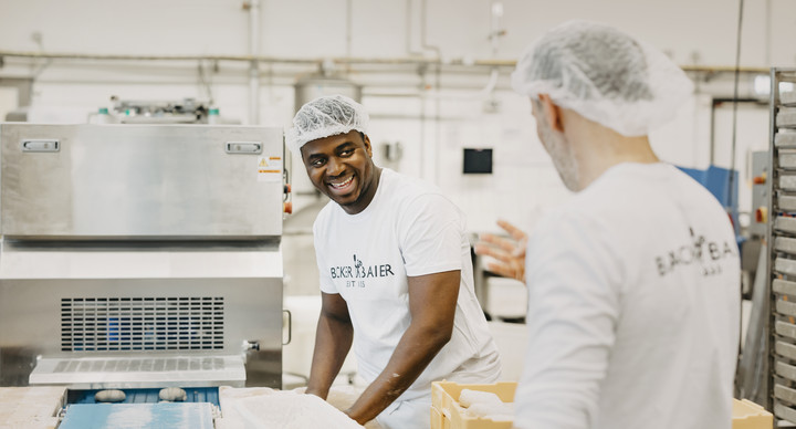 Zwei Männer arbeiten in einer Bäckerei. Einer lacht dem anderen zu, während er mit den Händen Backwaren bearbeitet. Im Hintergrund ist die Backstube zu sehen.
