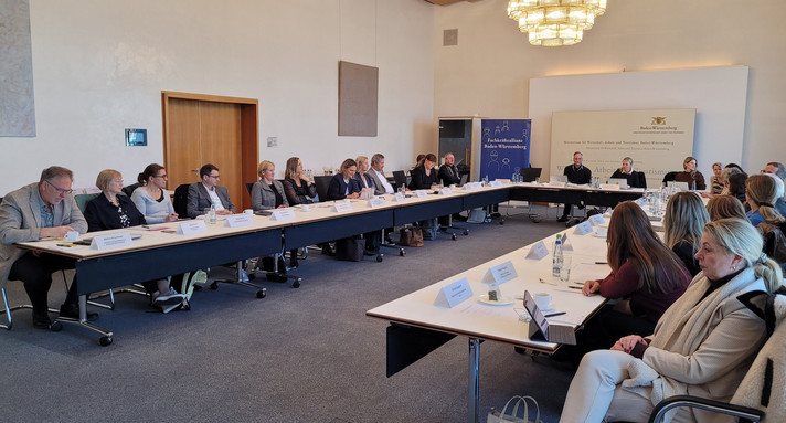 Auf dem Bild ist eine Gruppe von Menschen zu sehen, die an einem U-förmigen Tisch zusammensitzen. Vor Ihnen stehen Namensschilder.