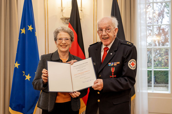 Staatsrätin Barbara Bosch (links) und Walter Adler (rechts) stehen vor Fahnen und zeigen die Verleihungsurkunde.