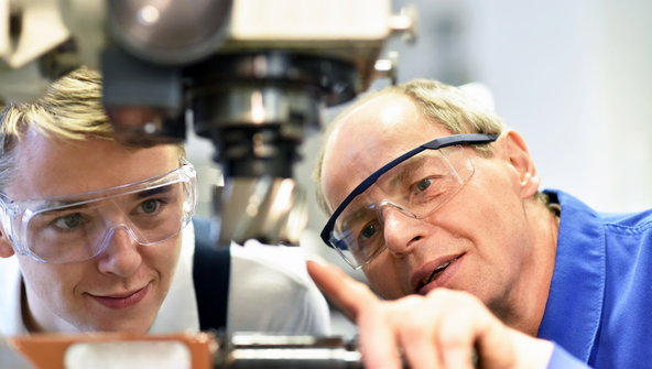 Ein Mann mit einer Schutzbrille zeigt auf eine Maschine. Neben ihm ein junger Mann ebenfalls mit einer Schutzbrille der darauf schaut.