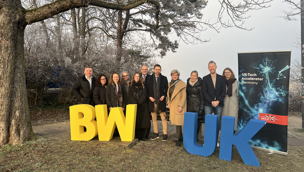 Eine Gruppe Menschen steht auf einer Wiese. Im Hintergrund ist ein Baum zu sehen. Vor der Gruppe stehen gelbe und blaue Buchstaben: BW und UK.